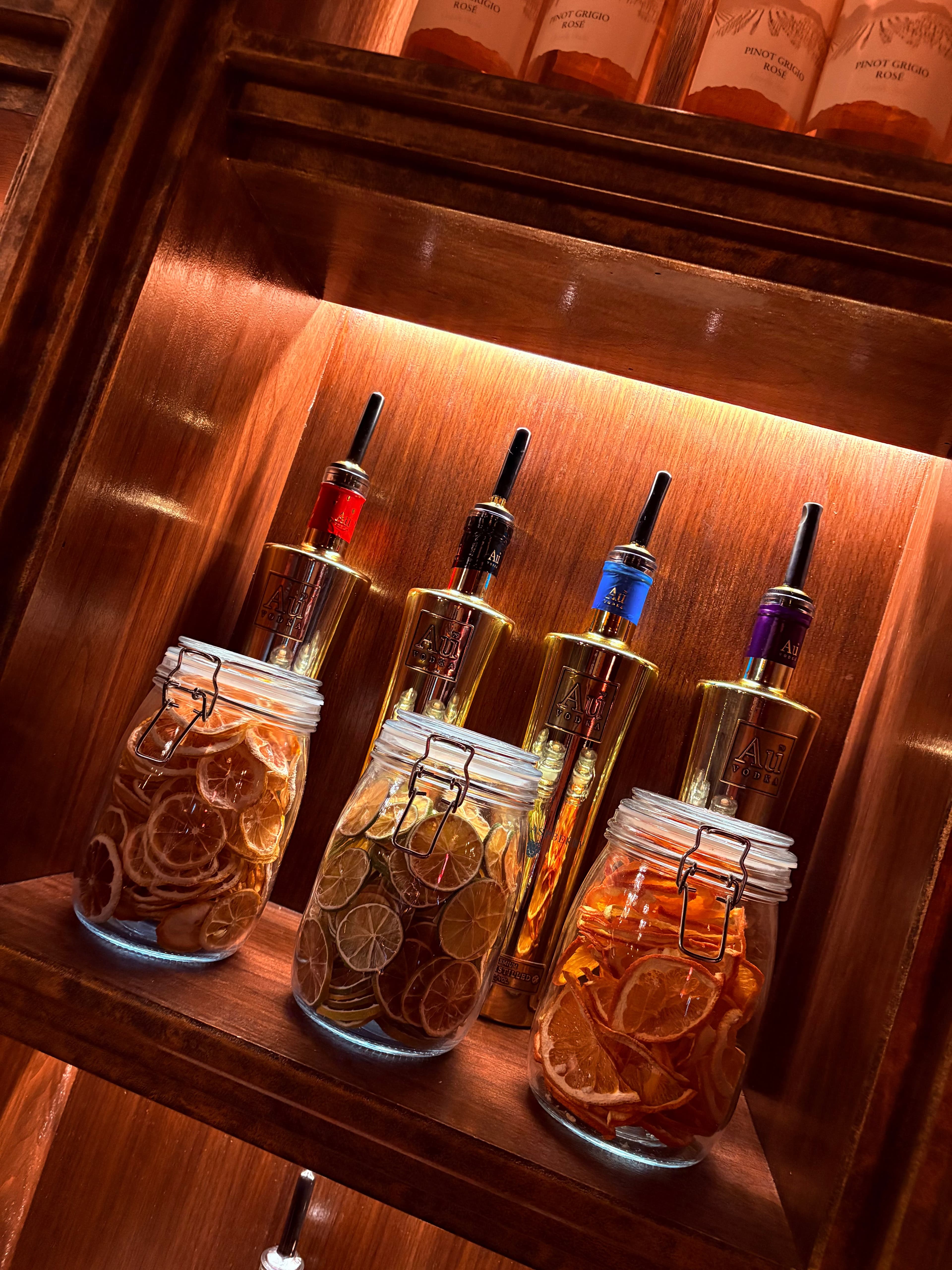 Au Vodka bottles with dried citrus garnish jars on backlit wooden shelf